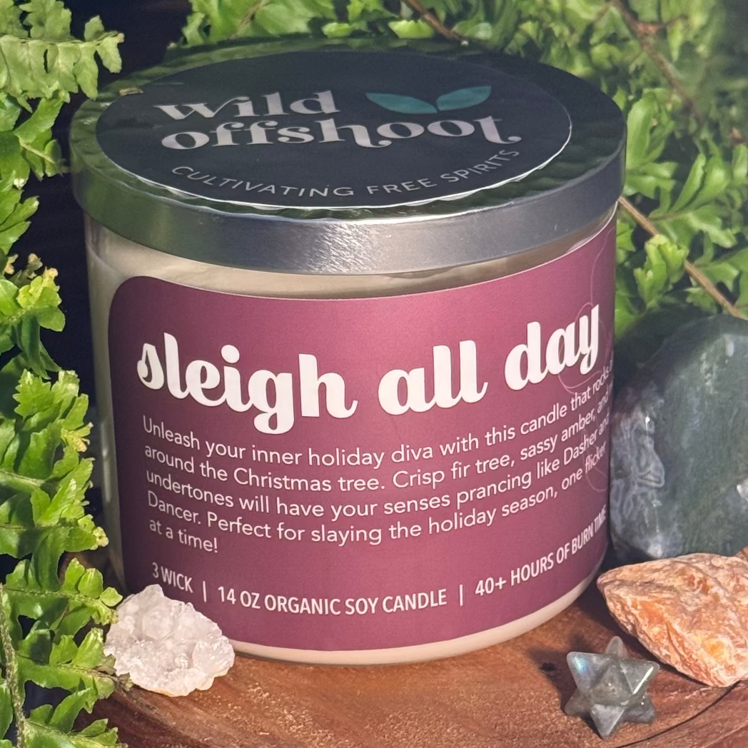 Container candle labeled 'sleigh all day' by Wild Offshoot with natural elements around it.