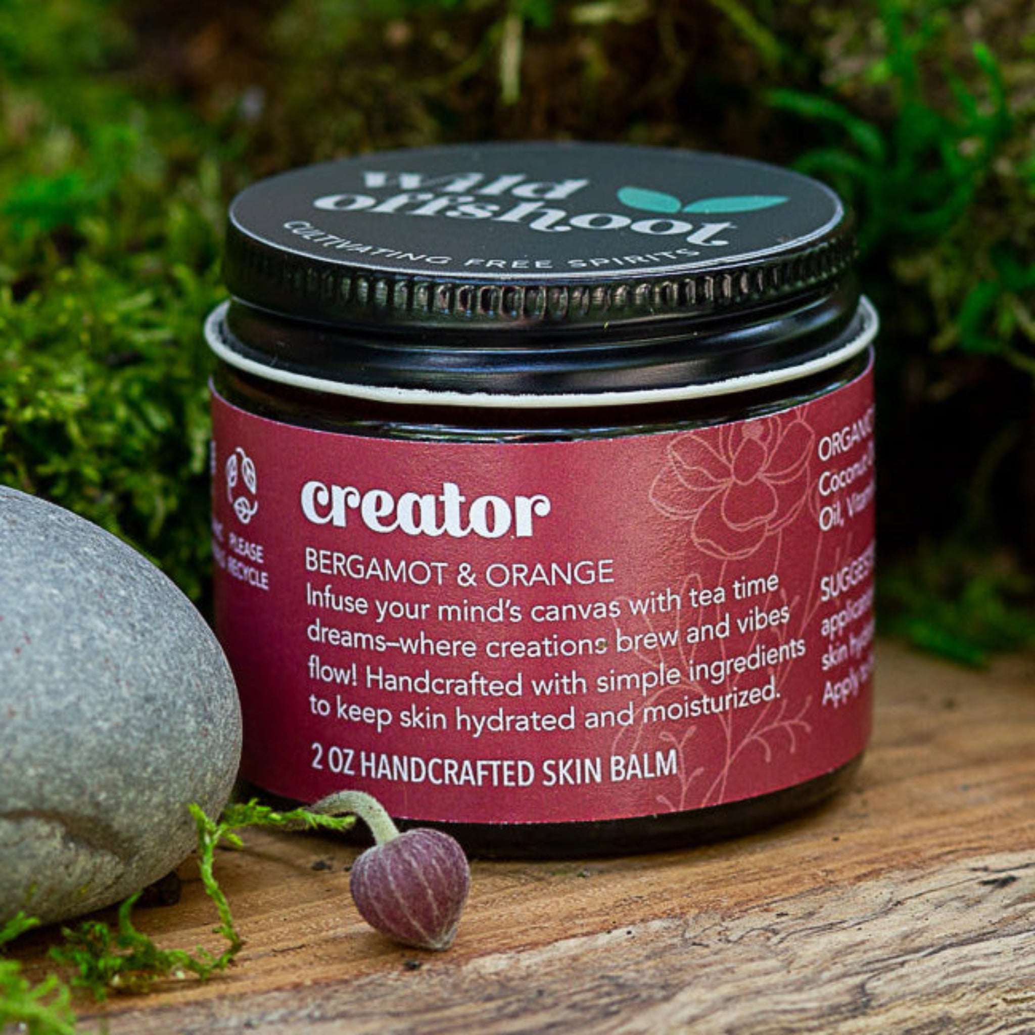 A 2 oz jar of Wild Offshoot Creator Body Balm with organic bergamot and orange, placed on a wooden surface with a rock and a flower nearby.