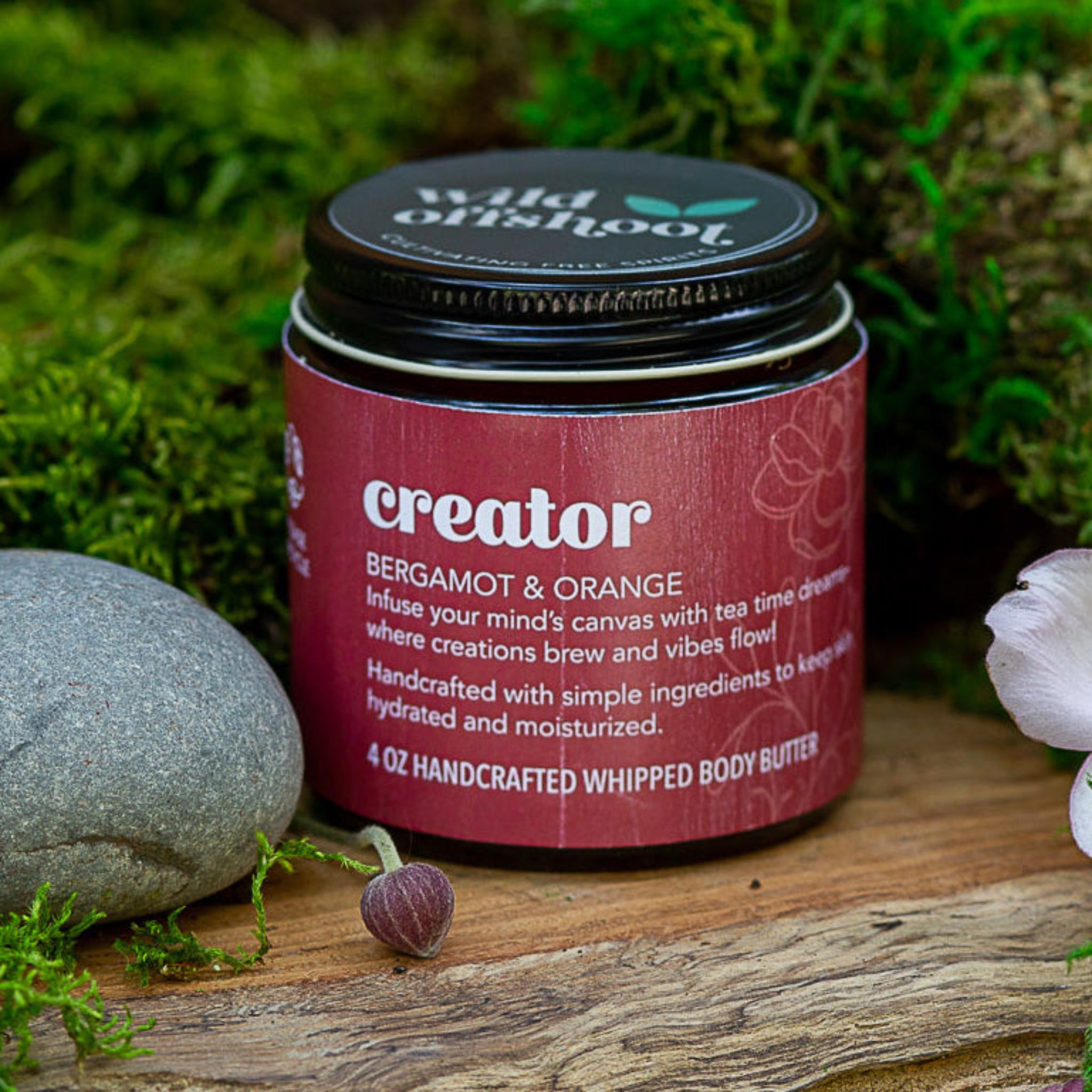 A 4 oz jar of Wild Offshoot Creator Body Balm with organic bergamot and orange, placed on a wooden surface with a rock and a flower nearby.