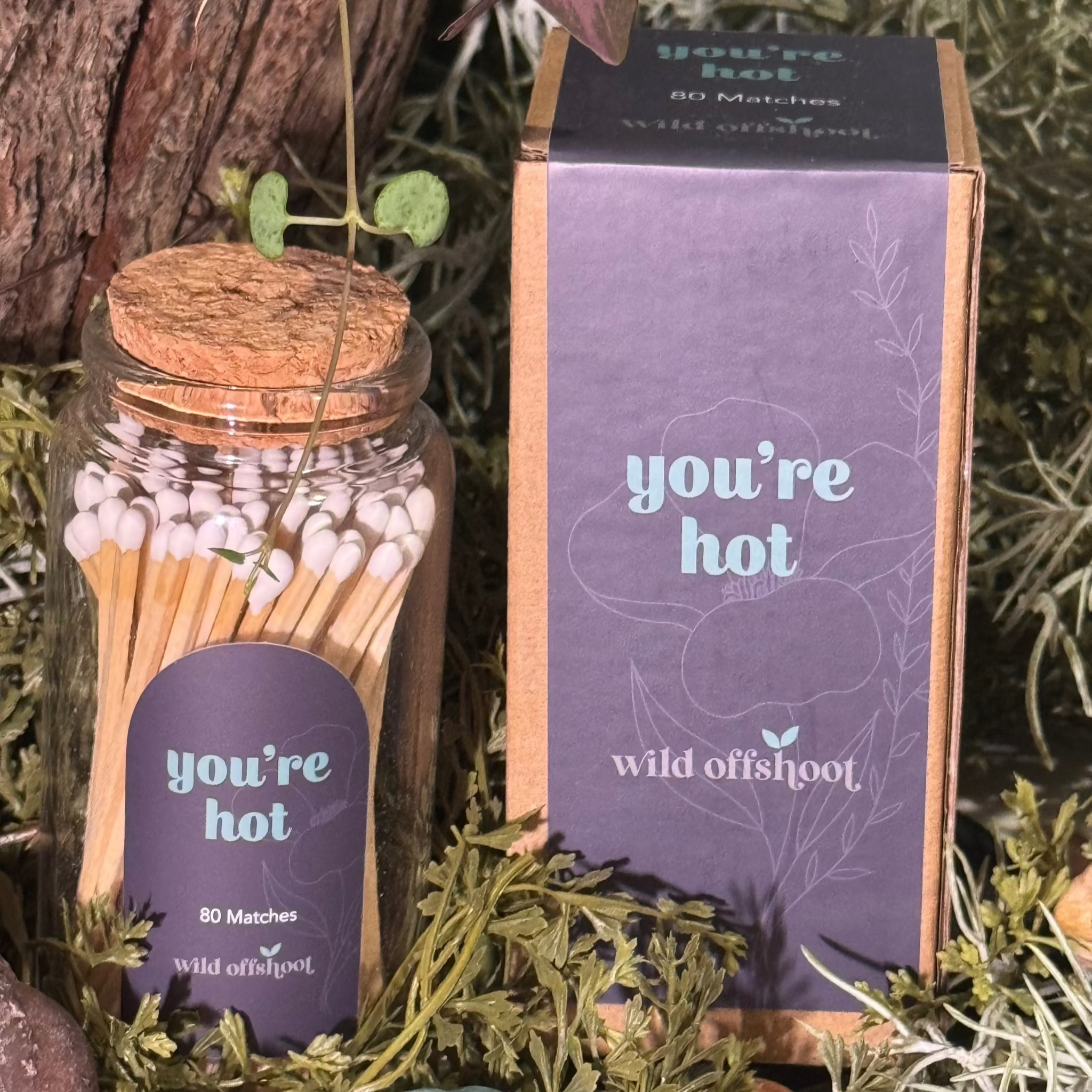 A glass jar with cork lid containing You're Hot Stick Matches, placed on a bed of artificial greenery next to a plant.