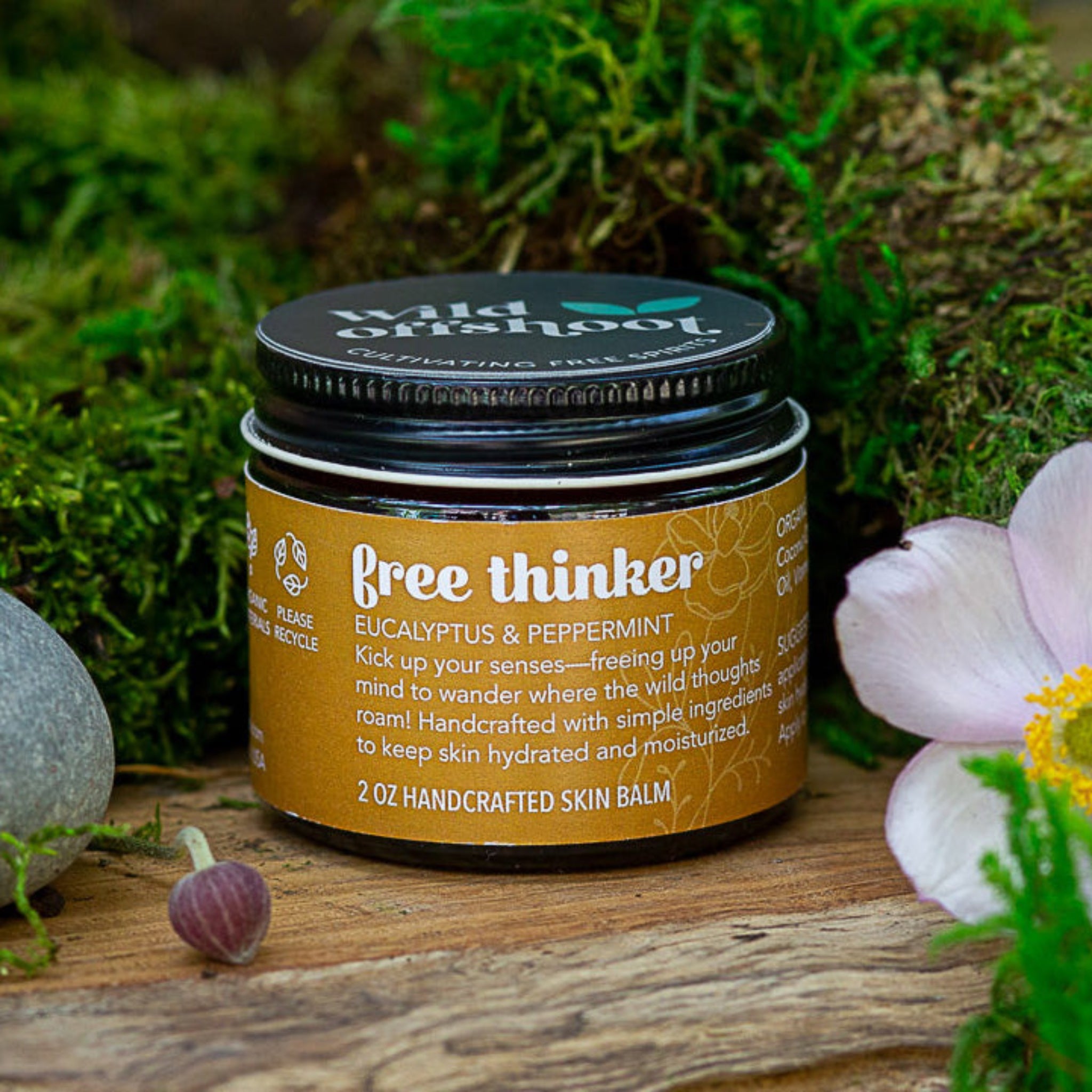 A 2oz jar of Wild Offshoot free thinker Body Balm with a label that includes the product name and a brief description, placed on a wooden surface surrounded by greenery and decorative stones.