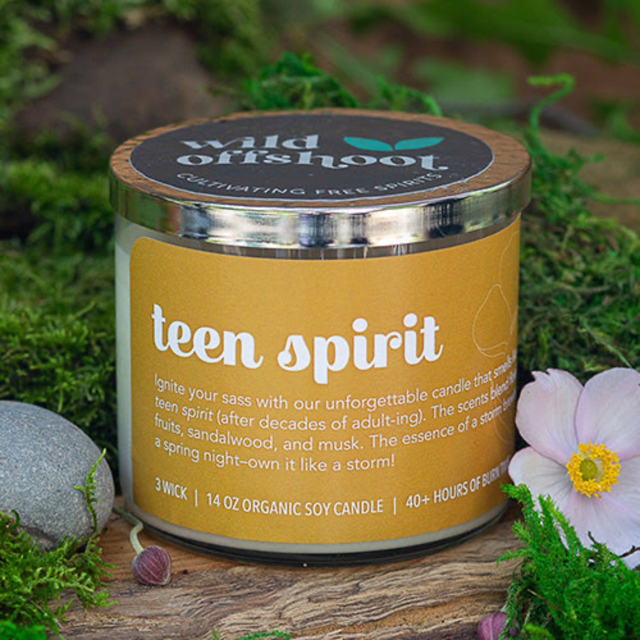 A gold glass 10 oz jar candle with a silver metal lid, labeled 'Teen Spirit' with text describing the scent profile. Three wicks and 40 hours of burn time.