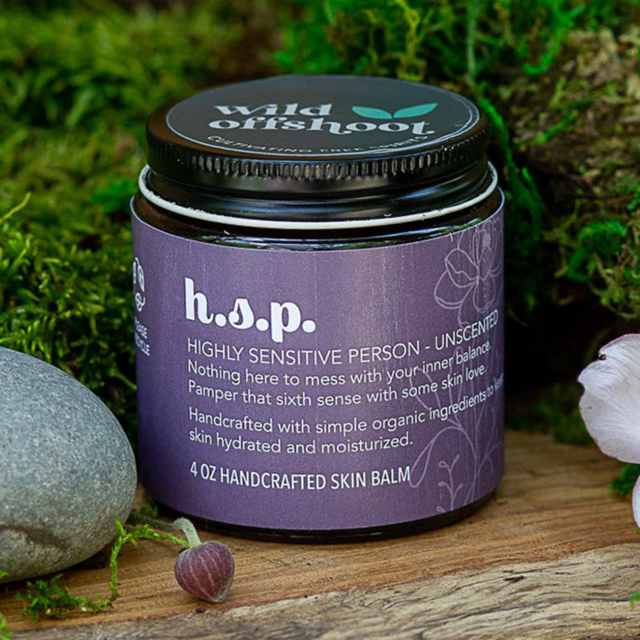 A 4oz jar of Wild Offshoot h.s.p. Body Balm with a label that includes the product name and a brief description, placed on a wooden surface surrounded by greenery and decorative stones.