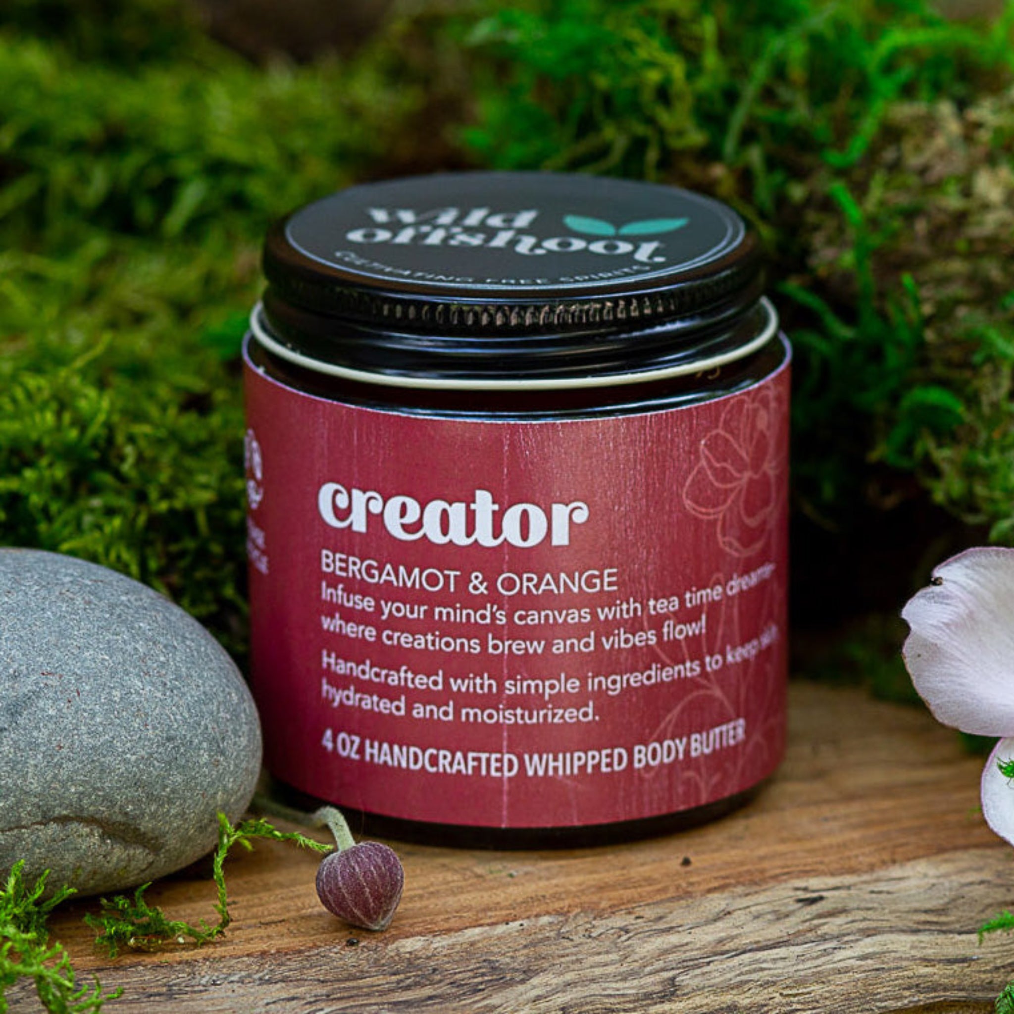 A 4 oz jar of Wild Offshoot Whipped Body Butter with the label 'Creator' showing a bergamot and orange scent. The jar is placed on a wooden surface with flowers and greenery in the background.