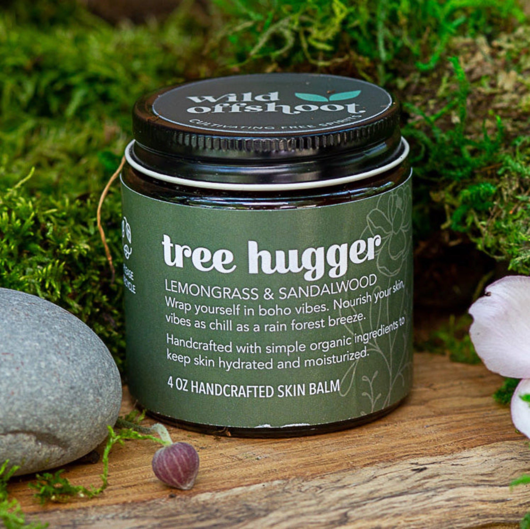 A 4oz jar of Wild Offshoot Flower Child Body Balm with a label that includes the product name and a brief description, placed on a wooden surface surrounded by greenery and decorative stones.