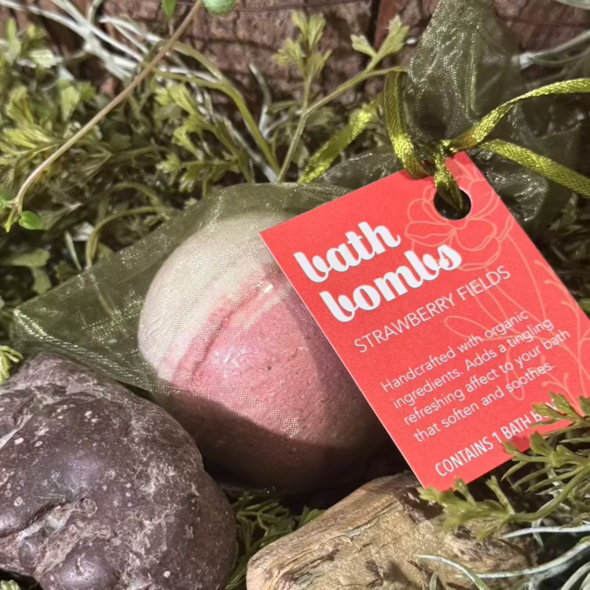 A bath bomb with a strawberry-themed label, surrounded by decorative greenery and rocks, with a tag showing the brand 'Wild Offshoot'.
