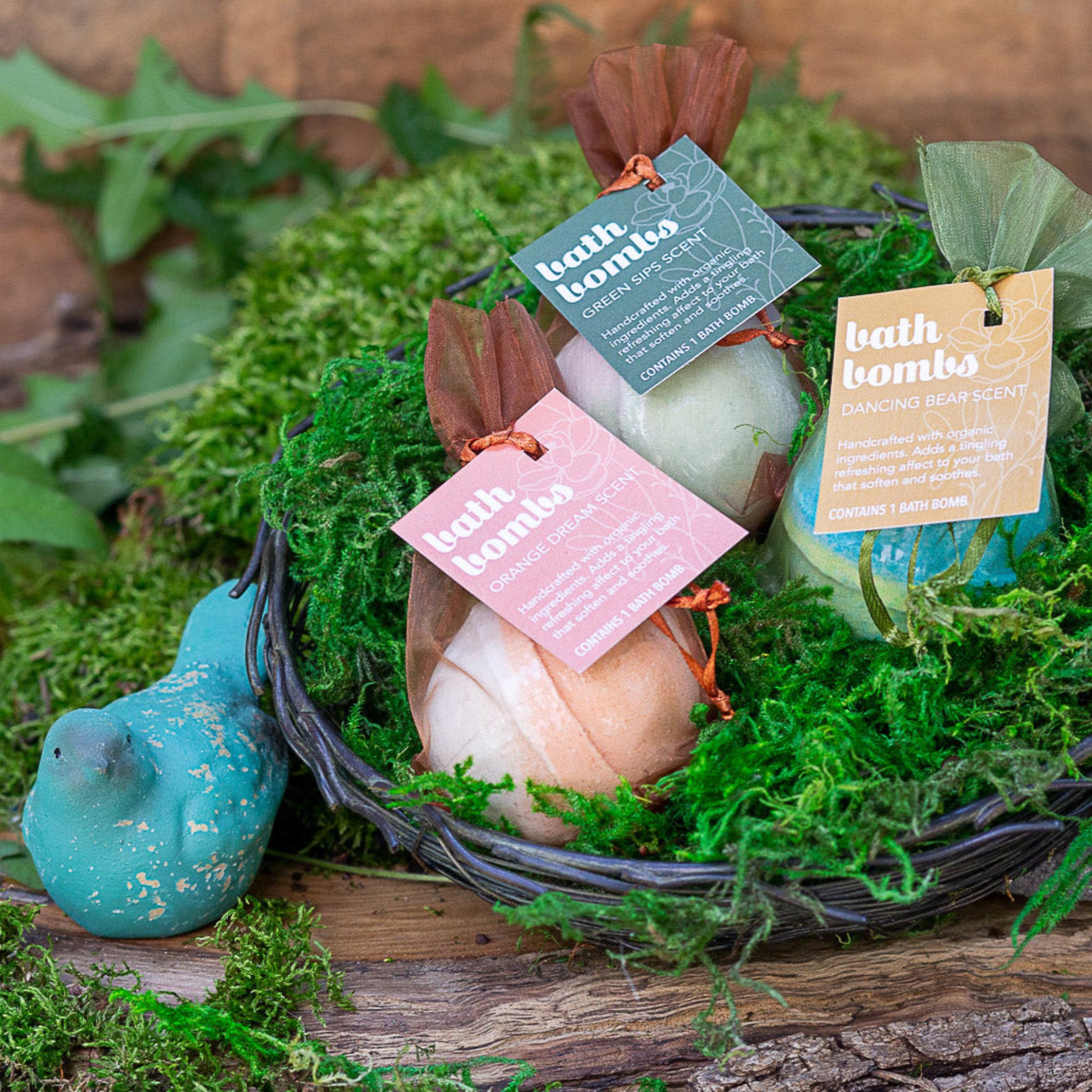 Three bath bombs in a basket, with each having a tag indicating different scent options such as 'Green Sips, 'Dancing Bear Scent,  and 'Orange Dream'. The bath bombs are placed on a bed of green moss-like material.