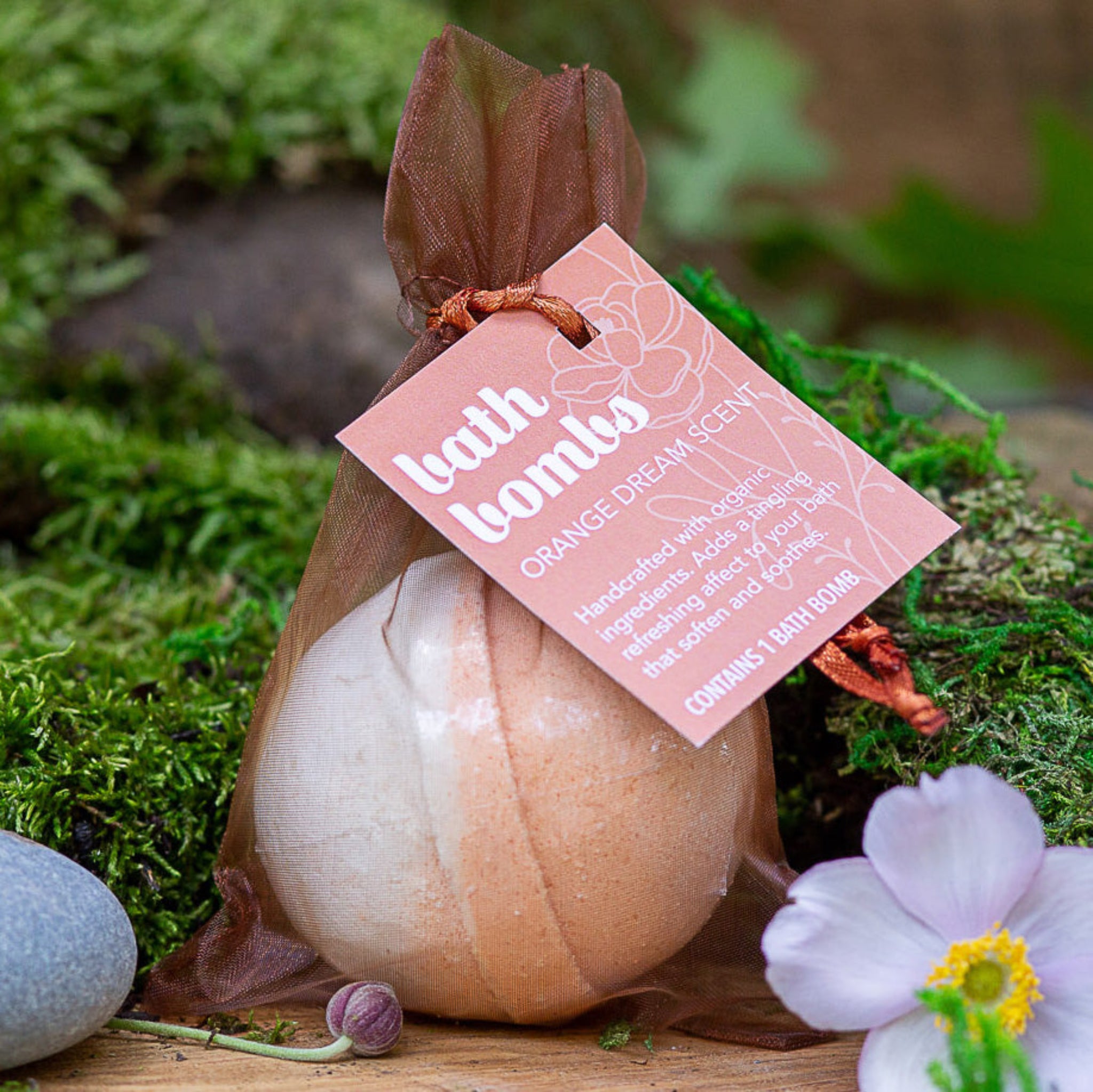 A packaged bath bomb with 'ORANGE DREAM' text on the label, placed on a wooden surface surrounded by green moss, a flower, and a rock.