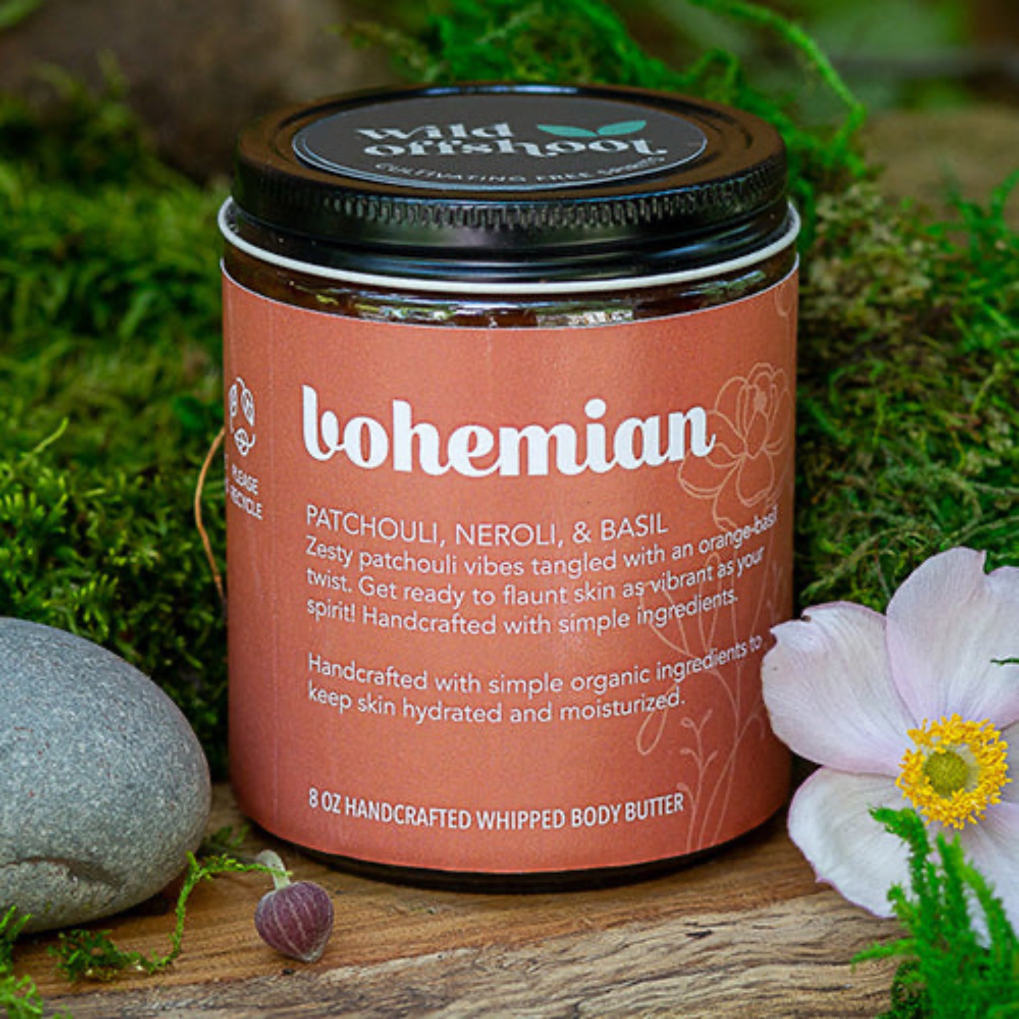 An 8oz jar of Wild Offshoot Whipped Body Butter with the label 'Bohemian' showing a patchouli, basil, and neroli scent. The jar is placed on a wooden surface with flowers and greenery in the background.
