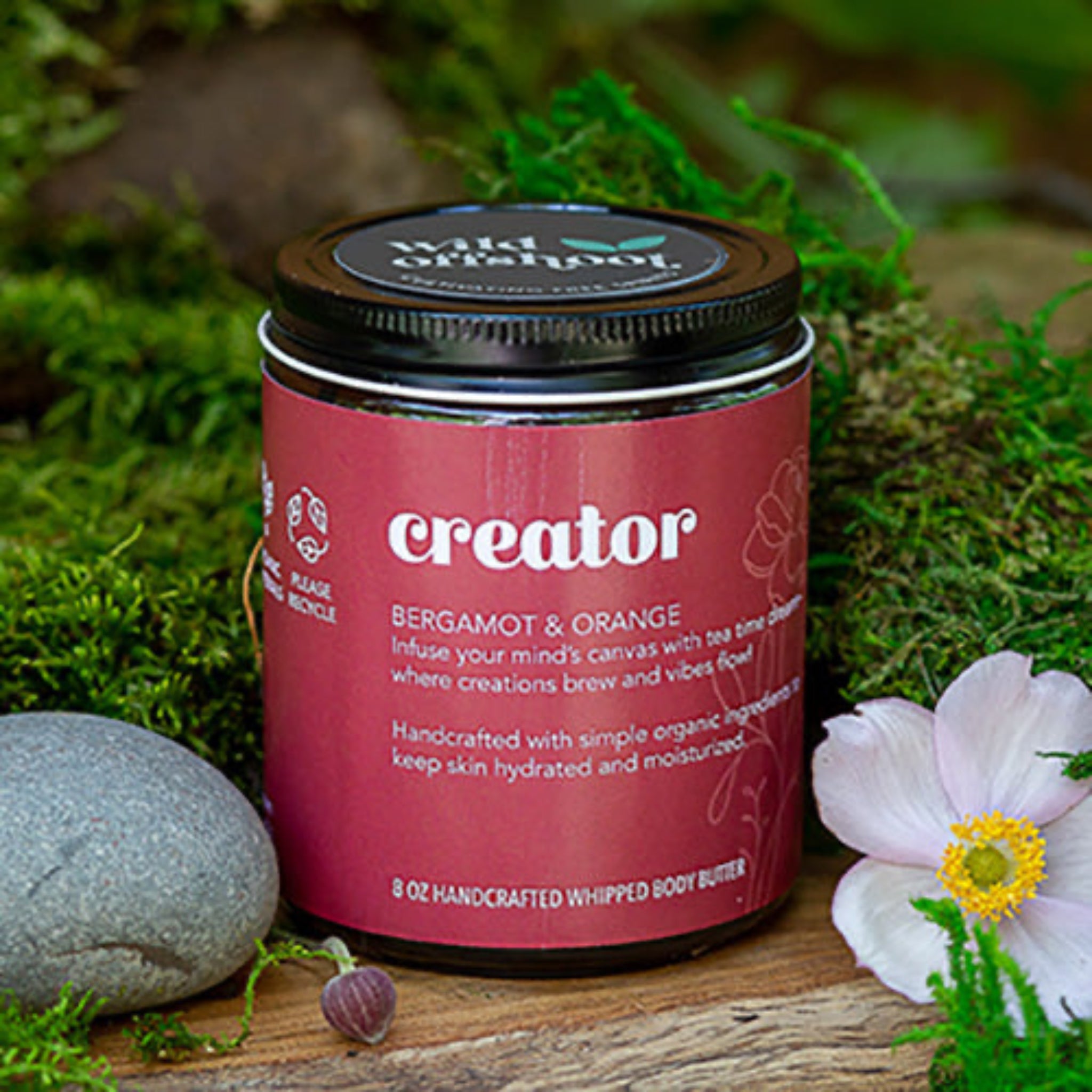 An 8 oz  jar of Wild Offshoot Whipped Body Butter with the label 'Creator' showing a bergamot and orange scent. The jar is placed on a wooden surface with flowers and greenery in the background.