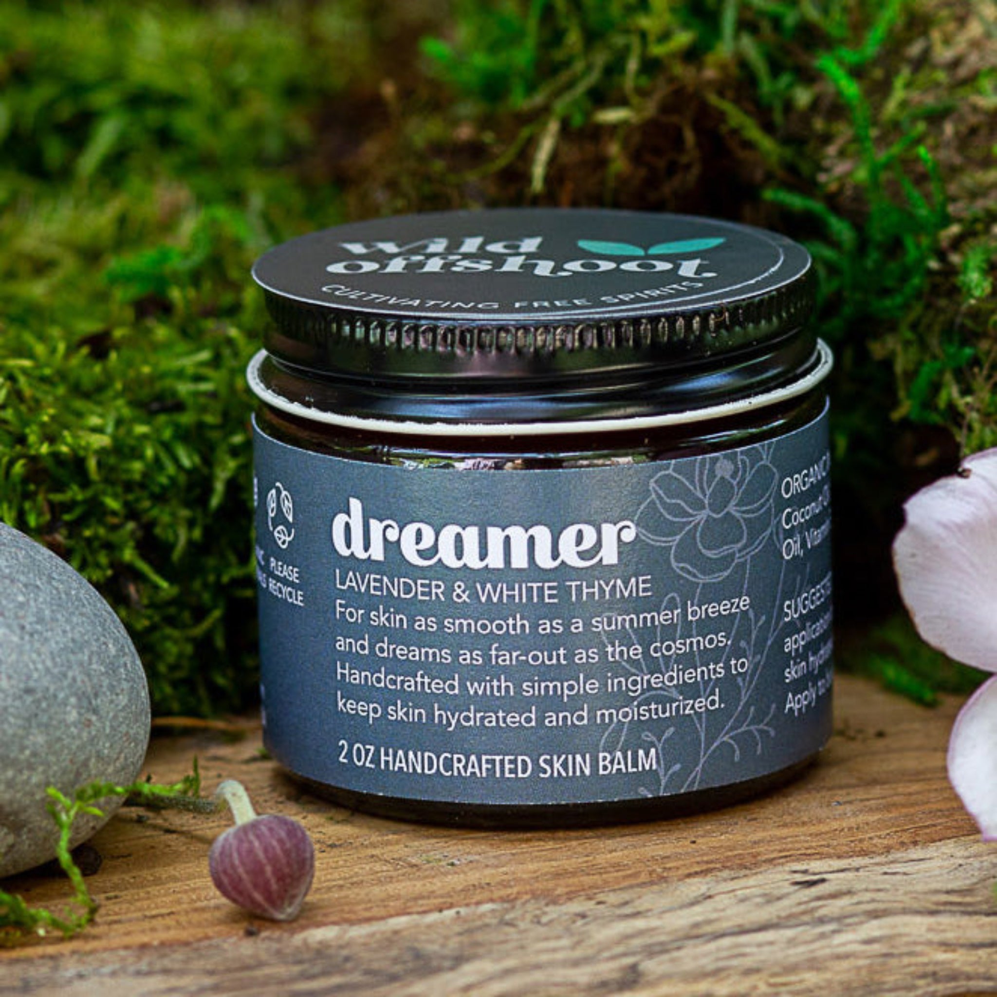 A 2 oz jar of Wild Offshoot Dreamer Body Balm with lavender and white thyme, placed on a wooden surface surrounded by greenery and decorative stones.