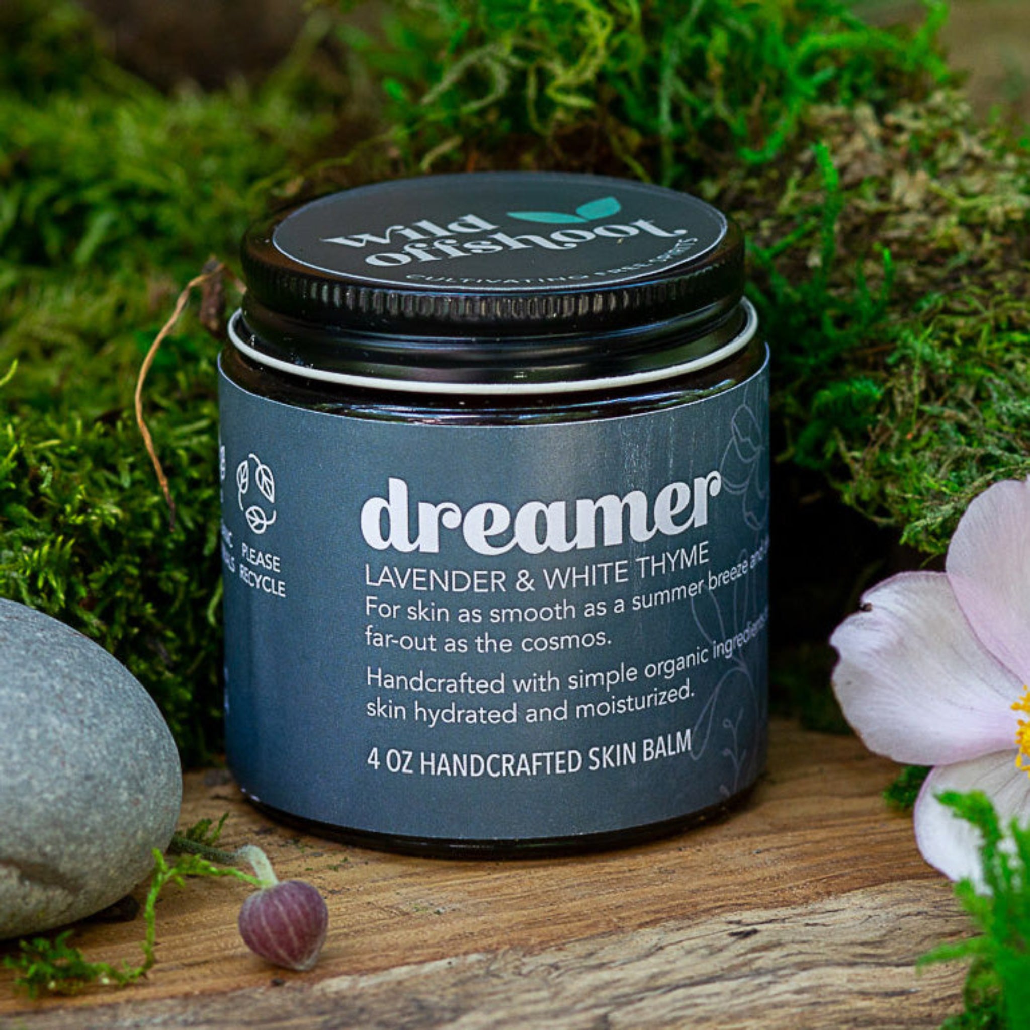 A 4 oz jar of Wild Offshoot Dreamer Body Balm with lavender and white thyme, placed on a wooden surface surrounded by greenery and decorative stones.