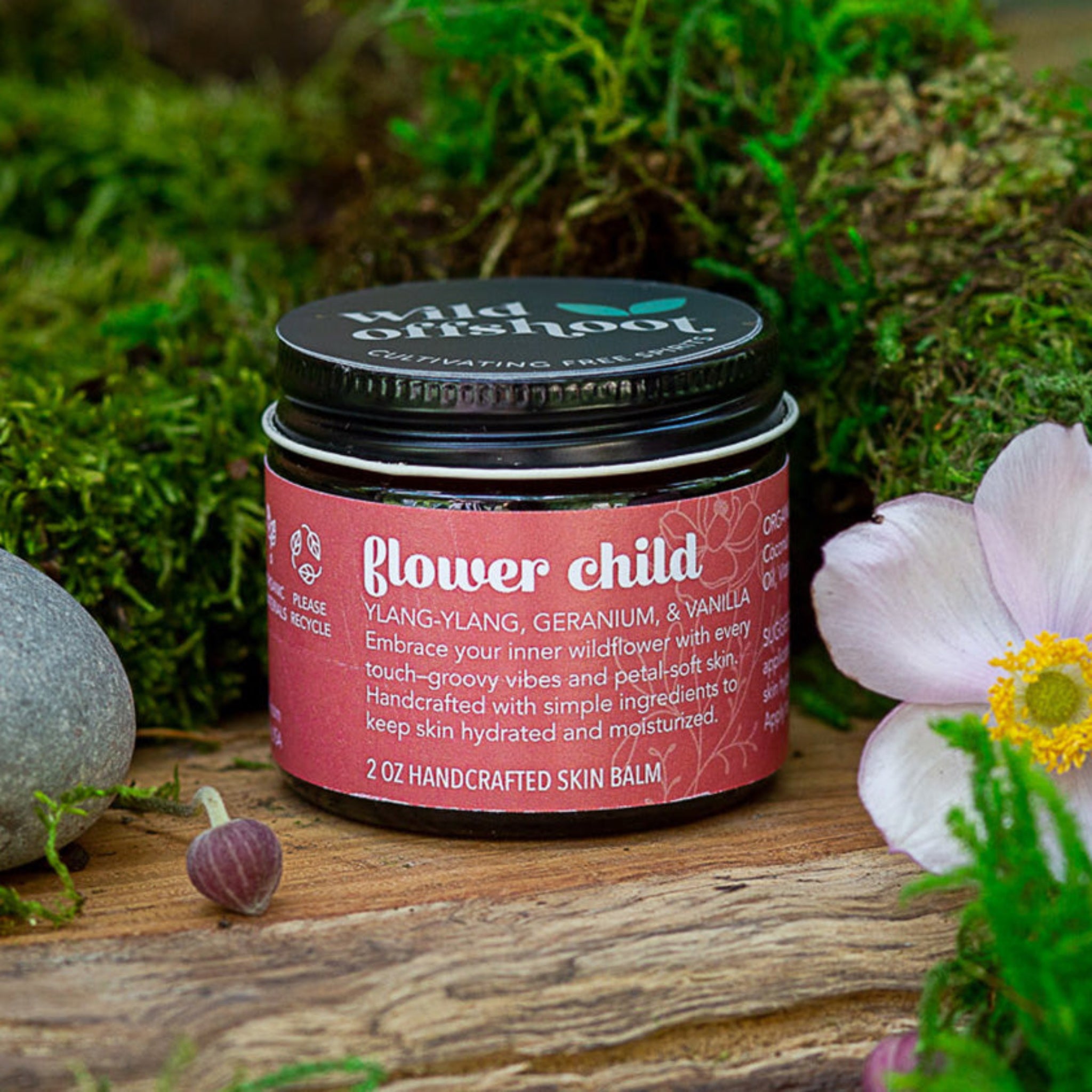 A 2oz jar of Wild Offshoot Flower Child Body Balm with a label that includes the product name and a brief description, placed on a wooden surface surrounded by greenery and decorative stones.