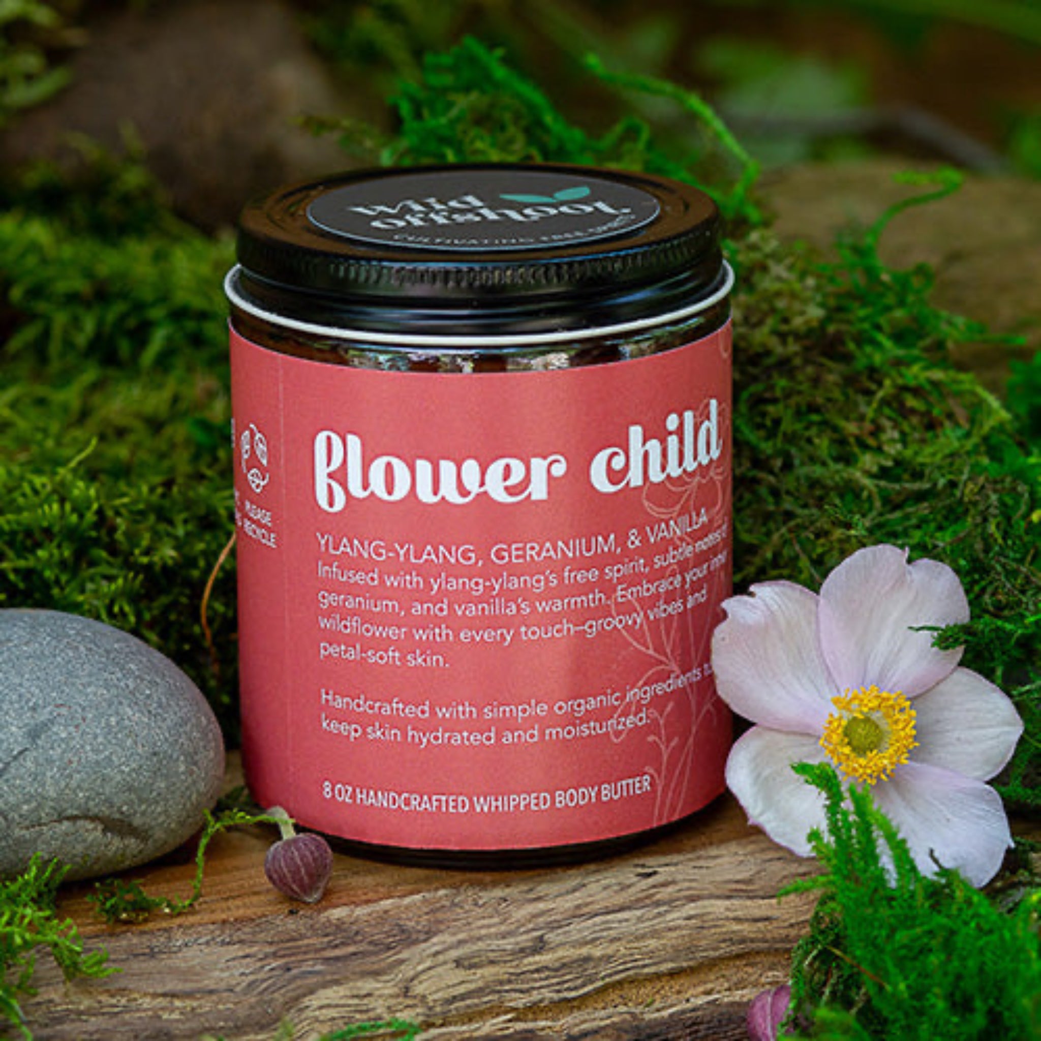 A 8 oz jar of Wild Offshoot Whipped Body Butter with the label 'flower child' showing a ylang-ylang, geranium, and vanilla scent. The jar is placed on a wooden surface with flowers and greenery in the background.