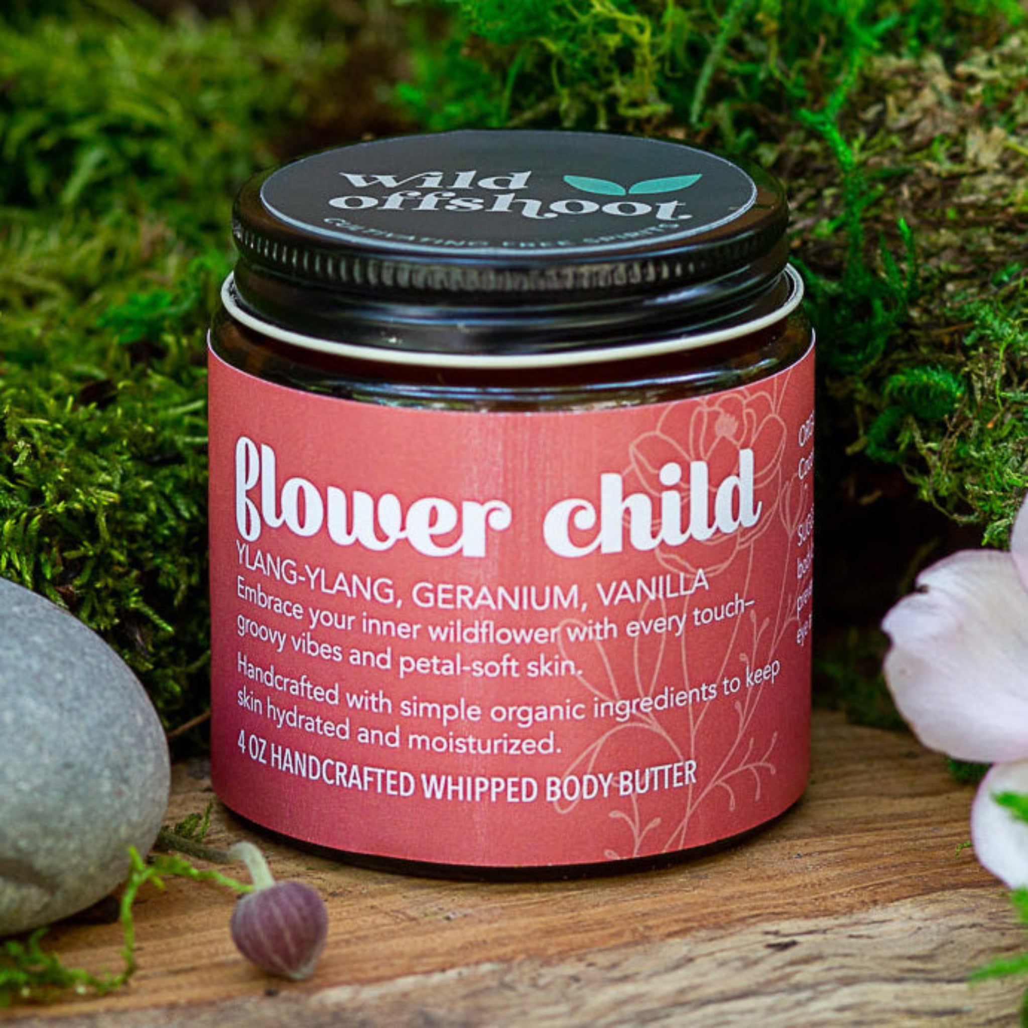 A 4 oz jar of Wild Offshoot Flower Child Whipped Body Butter  with ylang-ylang, geranium, and vanilla scent; placed on a wooden surface with decorative stones and flowers around it.