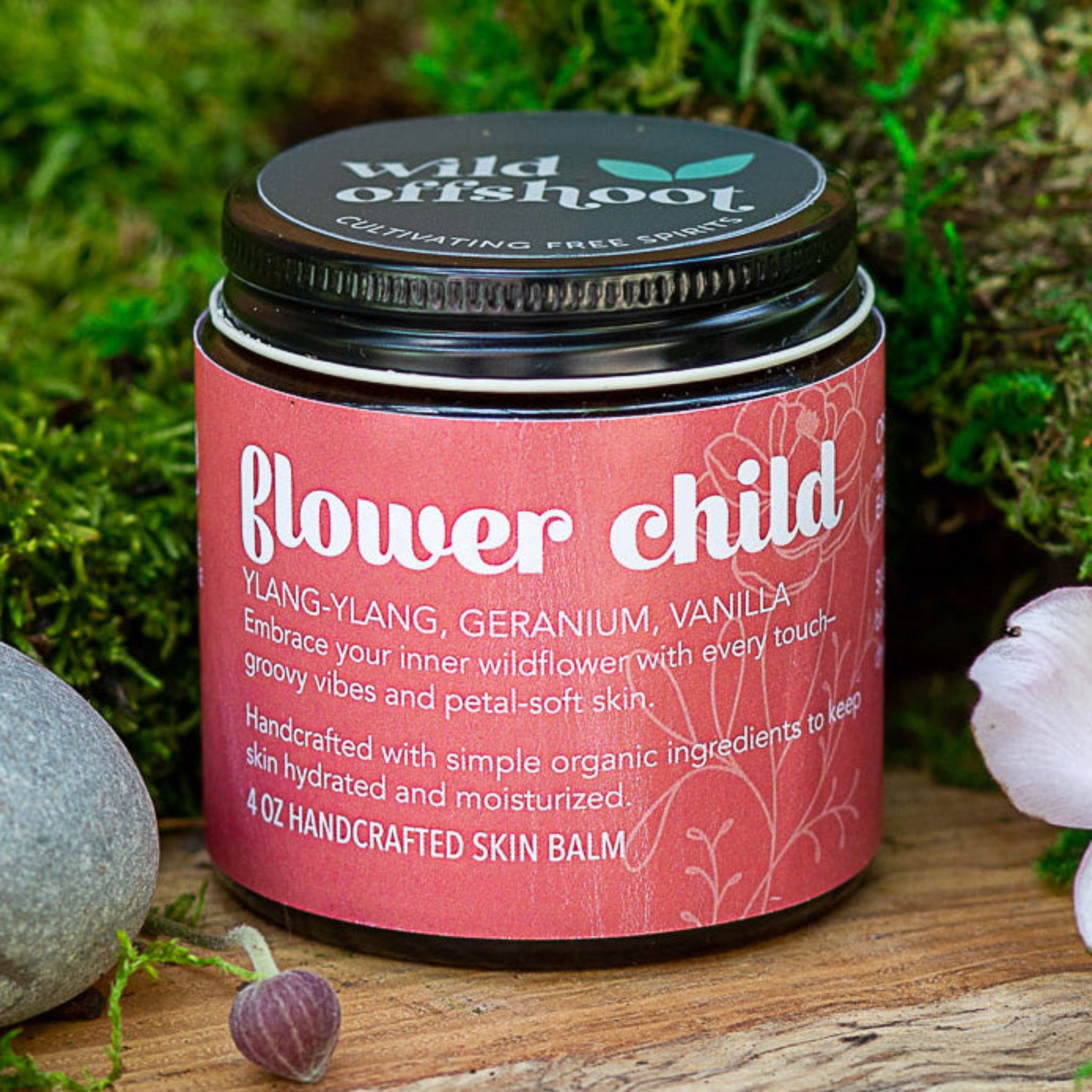 A 4oz jar of Wild Offshoot Flower Child Body Balm with a label that includes the product name and a brief description, placed on a wooden surface surrounded by greenery and decorative stones.