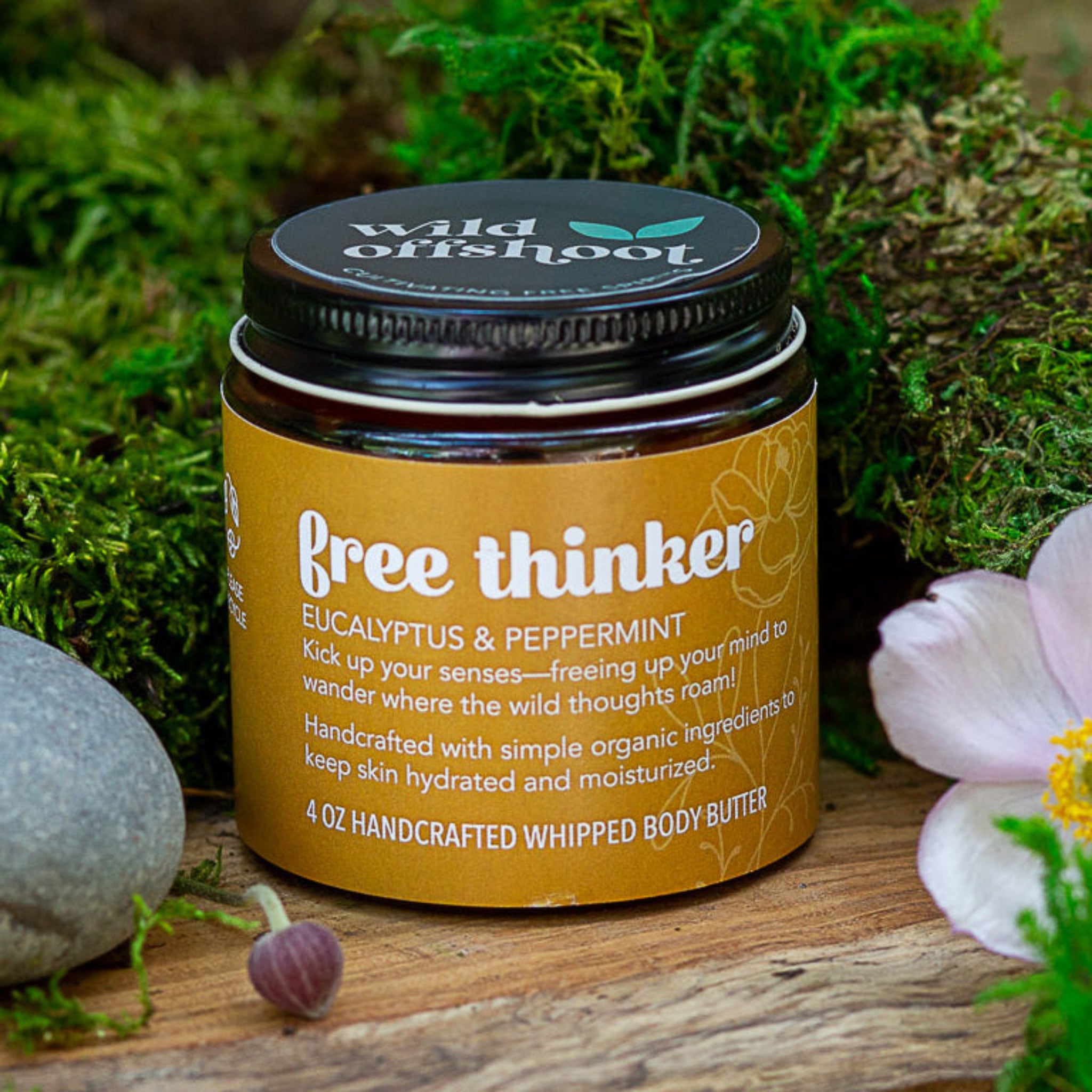 A 4 oz jar of Wild Offshoot Free Thinker Whipped Body Butter with organic eucalyptus and peppermint, placed on a wooden surface surrounded by greenery and decorative stones.