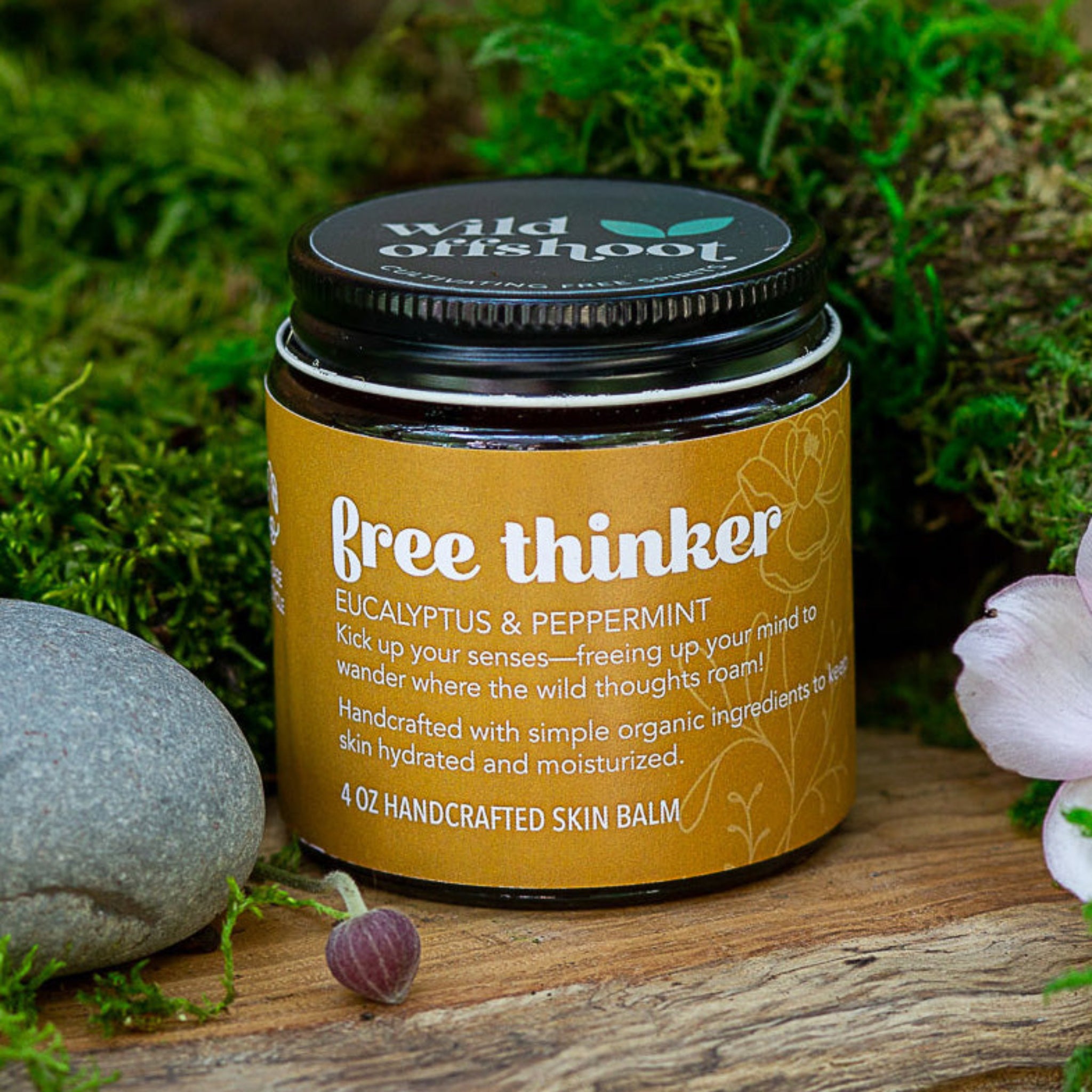 A 4oz jar of Wild Offshoot free thinker Body Balm with a label that includes the product name and a brief description, placed on a wooden surface surrounded by greenery and decorative stones.