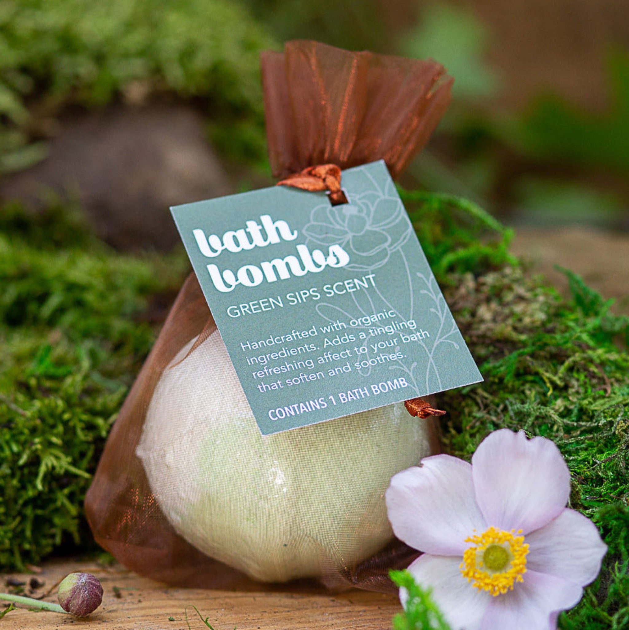 A packaged bath bomb labeled 'Green Sips'  The bath bomb is visible through a rust brown silk wrapping, set against a natural background with moss and flowers.