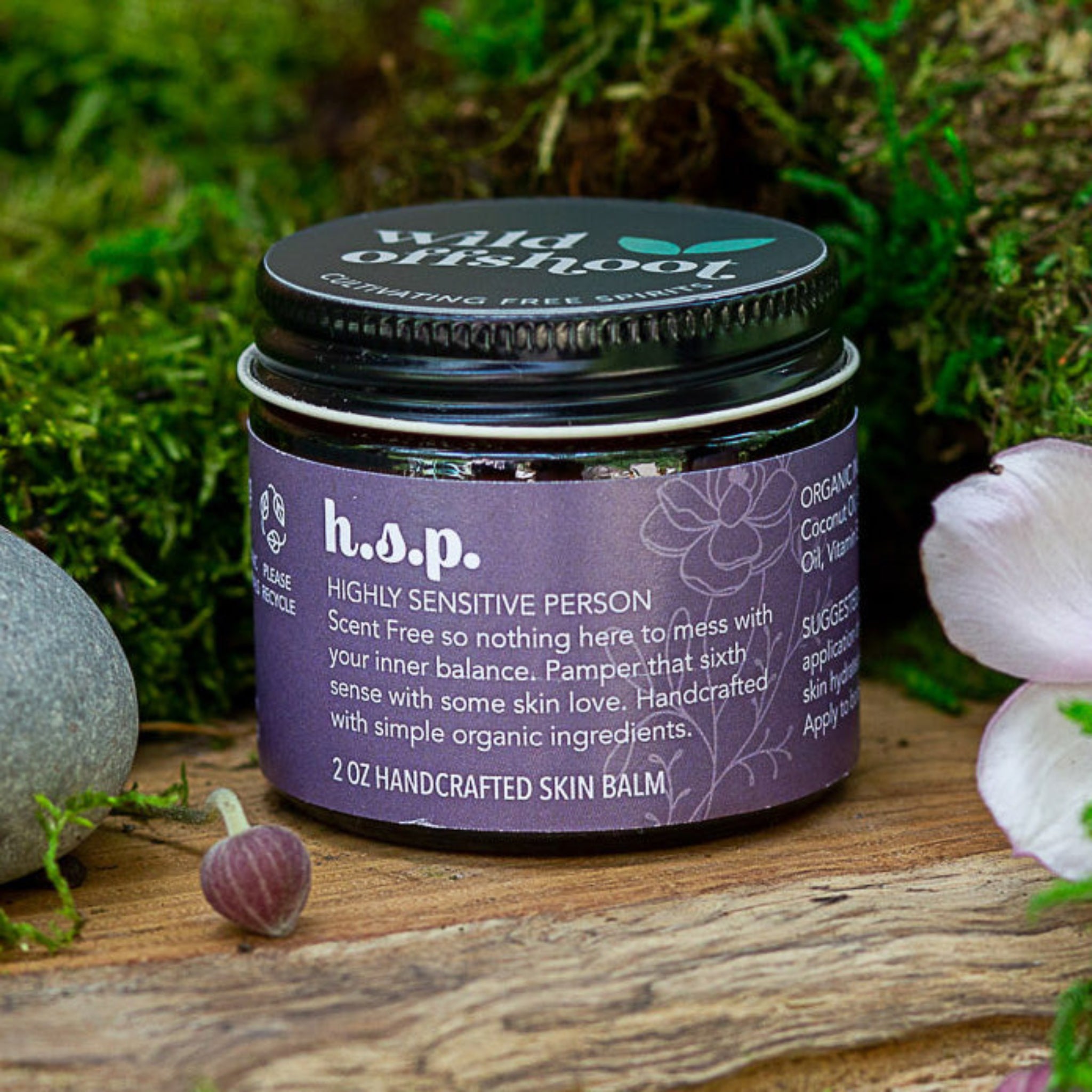 A 2oz jar of Wild Offshoot h.s.p. Body Balm with a label that includes the product name and a brief description, placed on a wooden surface surrounded by greenery and decorative stones.