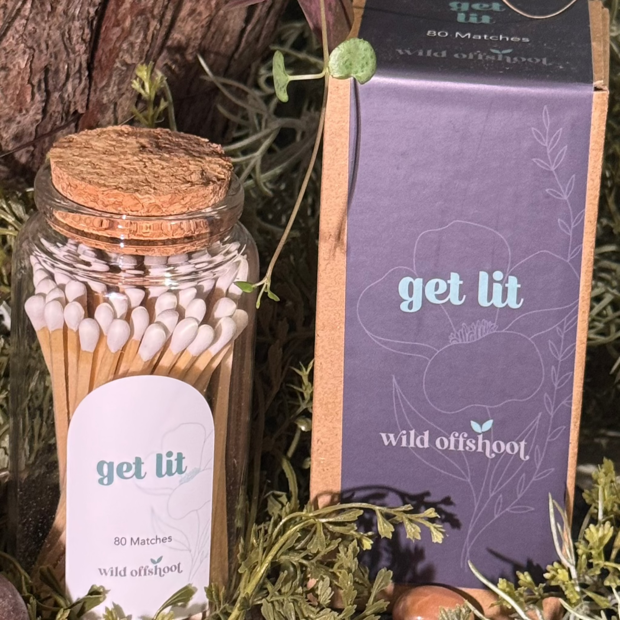A glass jar with cork lid containing 80 4-inch stick matches, labeled 'GET LIT Wild Offshoot', arranged on a bed of greenery and next to a box with 'Wild Offshoot' branding.
