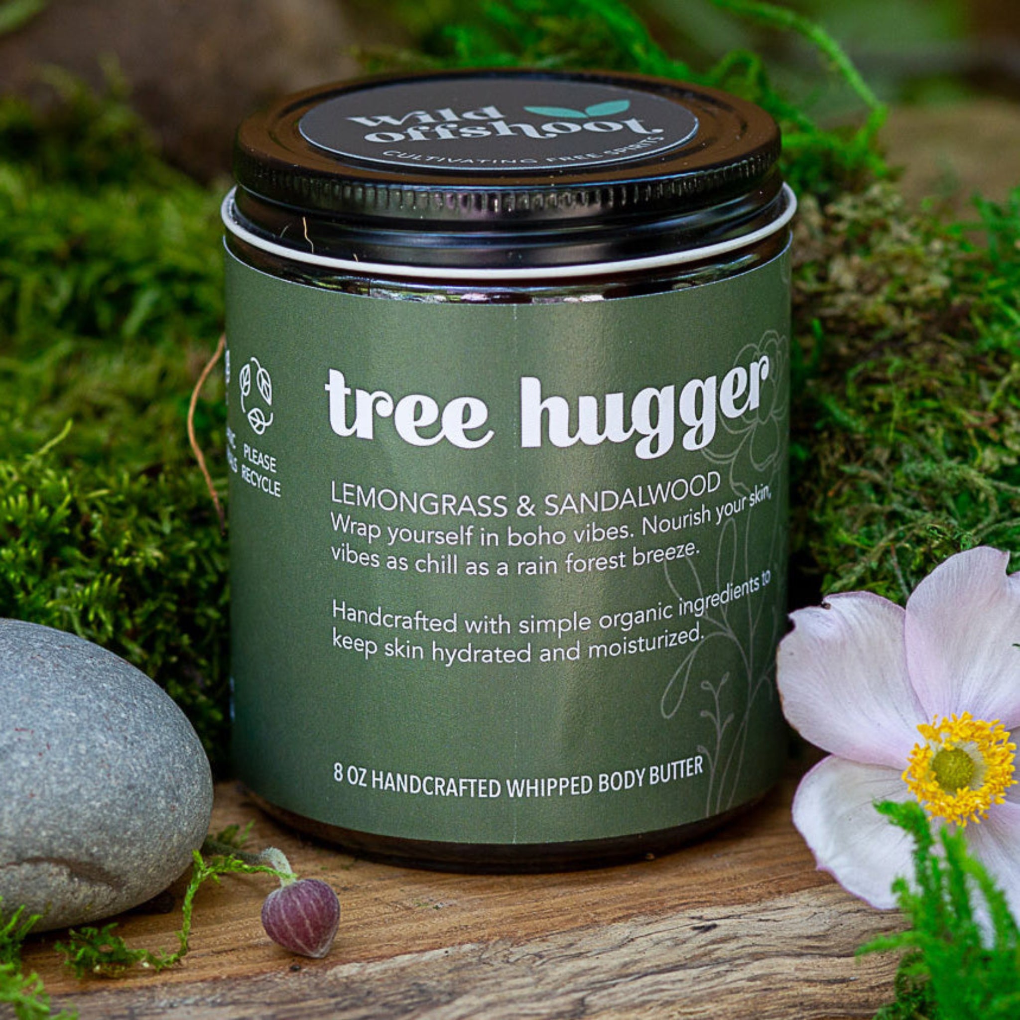An 8oz jar of Wild Offshoot Whipped Body Butter with a label indicating it is Tree Hugger with lemongrass and sandalwood scent - placed on a wooden surface with flowers and a rock in the background.
