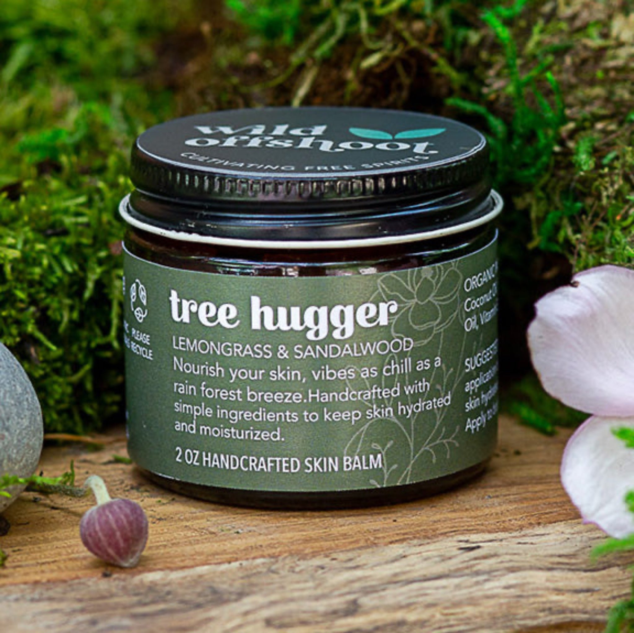 A 2oz jar of Wild Offshoot Tree Hugger Body Balm with a label that includes the product name and a brief description, placed on a wooden surface surrounded by greenery and decorative stones.