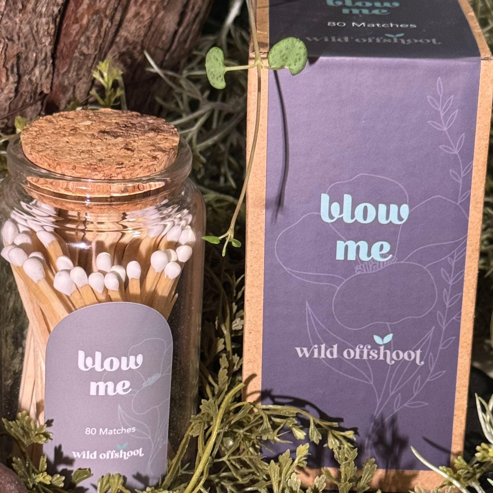 A glass jar with a cork lid containing 80 stick matches, labeled 'BLOW ME'. The jar is placed on a bed of decorative greenery with a cardboard box in the background.