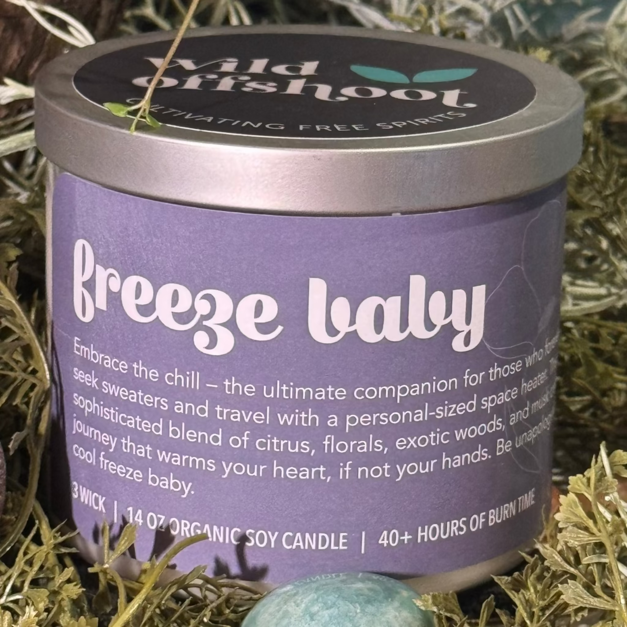 A purple candle labeled 'Freeze Baby' with a silver metal lid, placed on a table surrounded by greenery and a crystal.