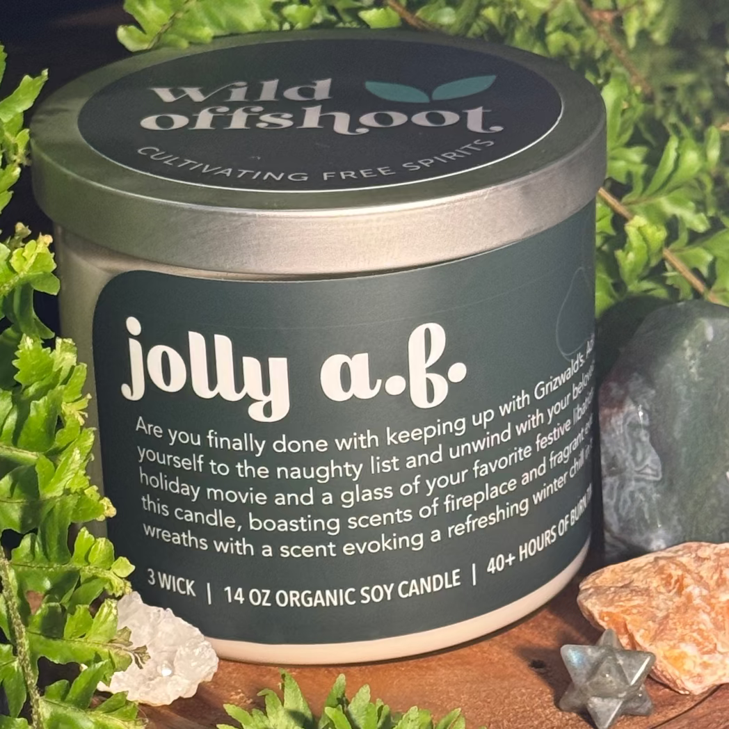 Container candle labeled 'jolly a.b.' with natural elements like plants and stones on a wooden surface.