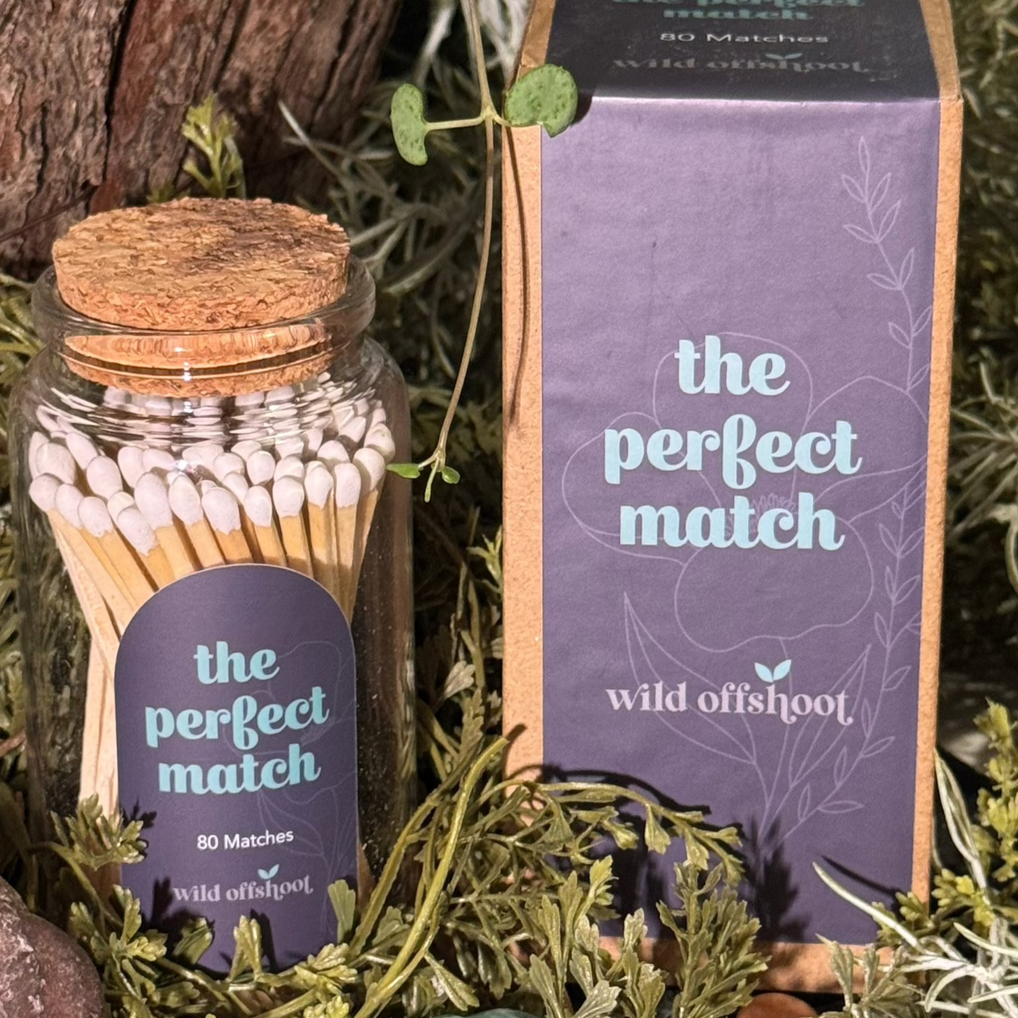 A glass jar with cork lid containing white stick matches, labeled 'The Perfect Match', with a box of the same product beside it, placed on a bed of greenery.