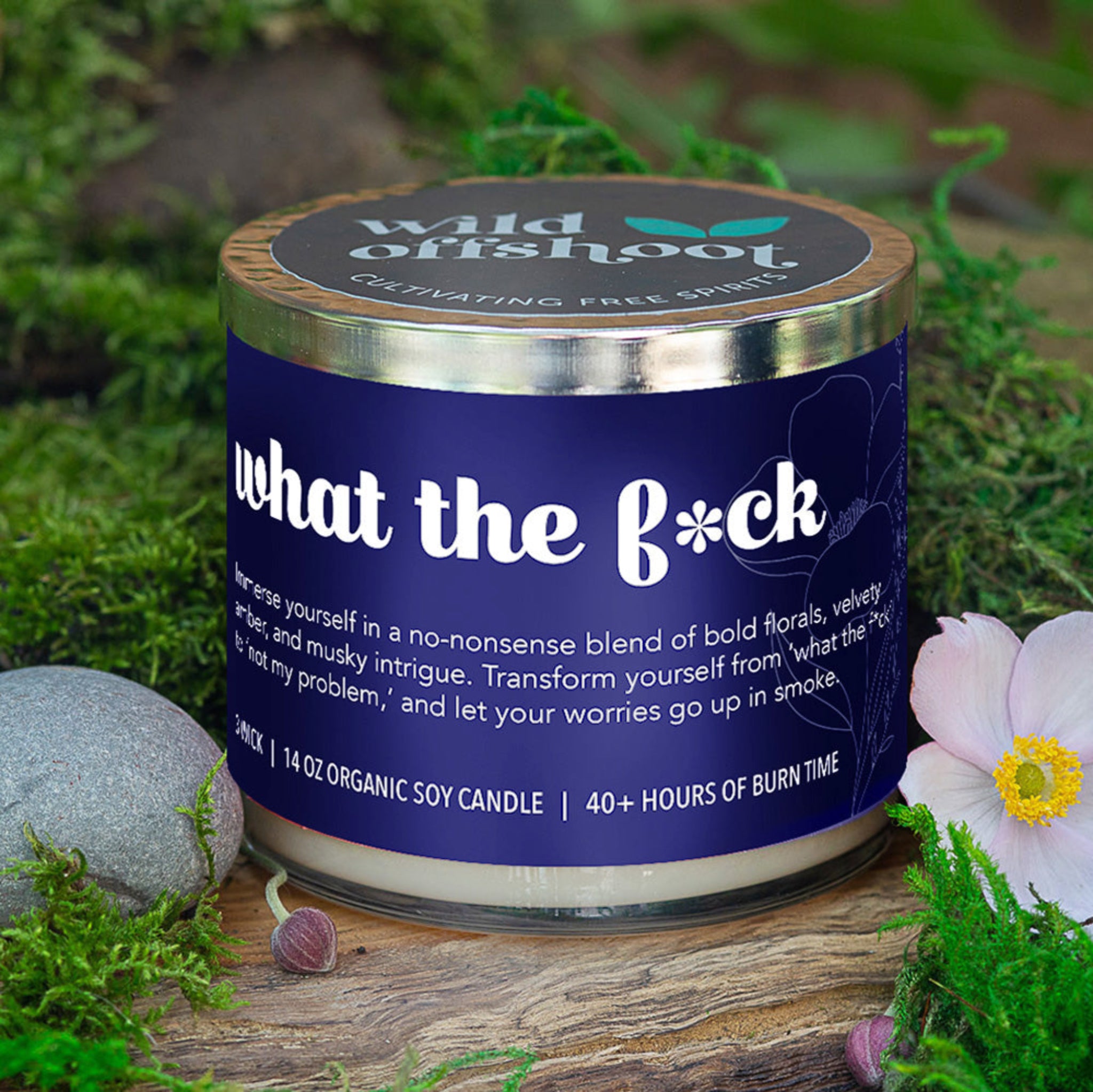 A blue glass jar candle with a silver metal lid, labeled 'WHAT THE F*CK' along with the brand 'Wild Offshoot'. The label also includes text describing the scent as a 'Bold Floral' and indicates the candle is 100% organic soy wax with 40 hours of burn time.